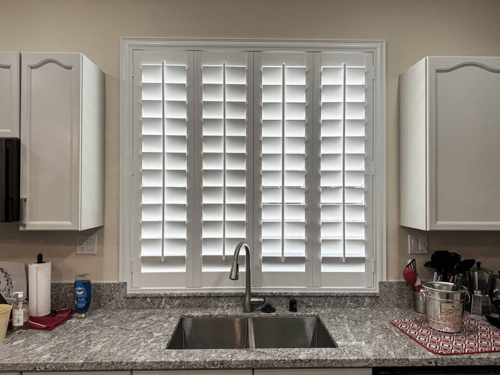 Four-panel white plantation shutters offer full privacy above a double stainless steel kitchen sink.