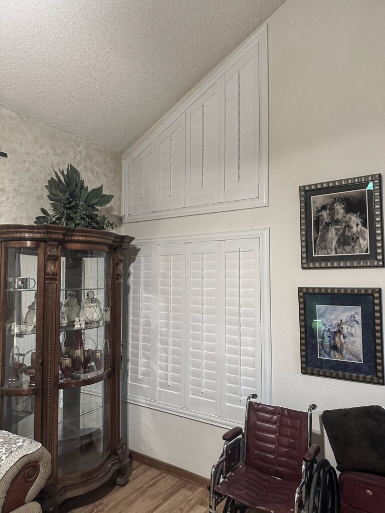 White custom-angled shutters cover a high-ceiling window above a display cabinet.