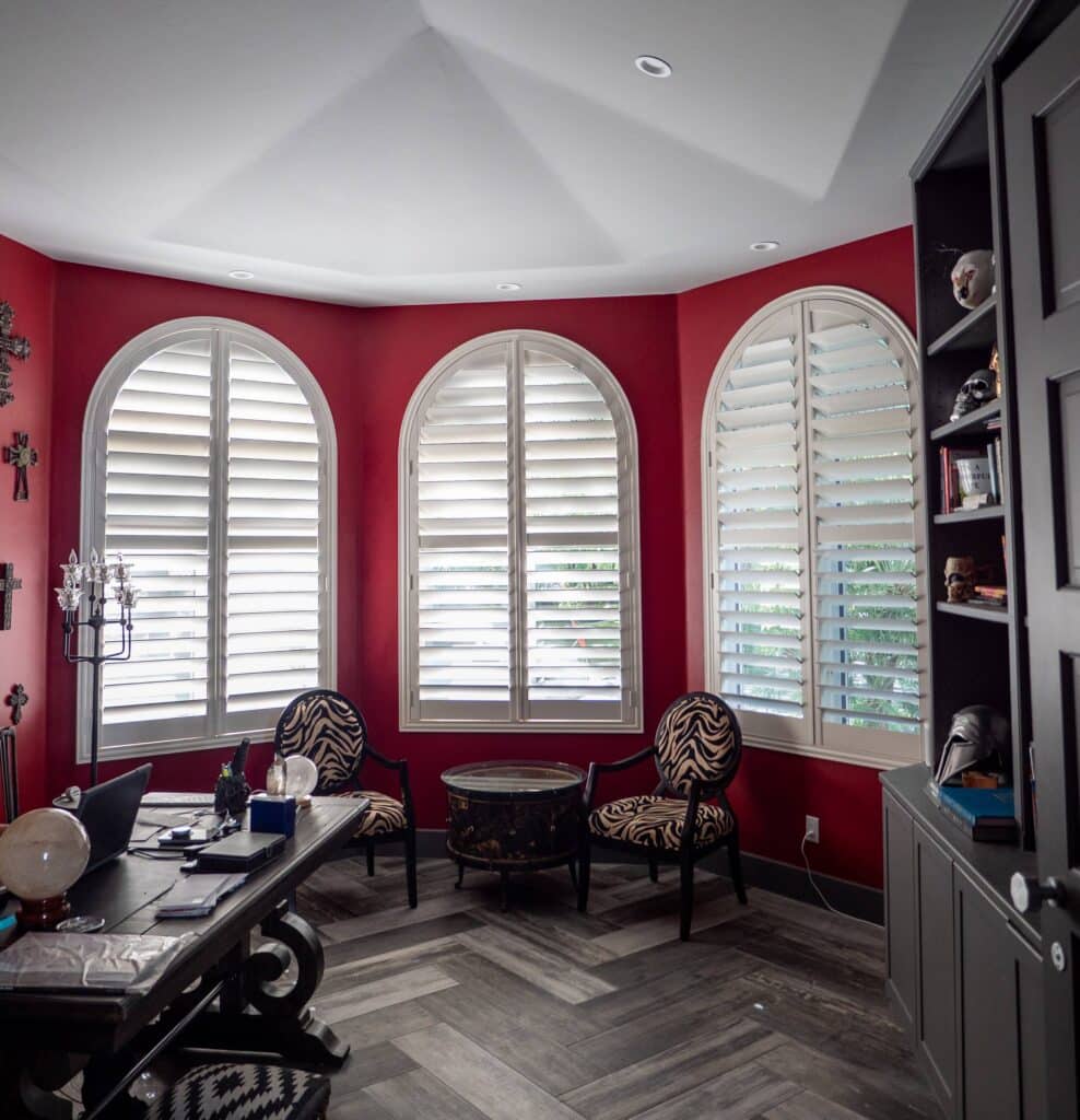Three arched white shutters stand out against bold red walls in a dramatically styled home office.