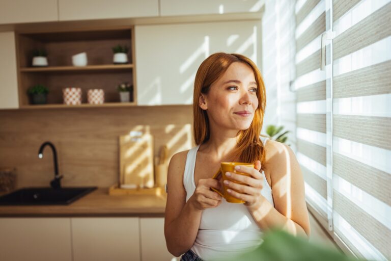 A woman holds a cup of coffee as she looks out the blinds in her kitchen.